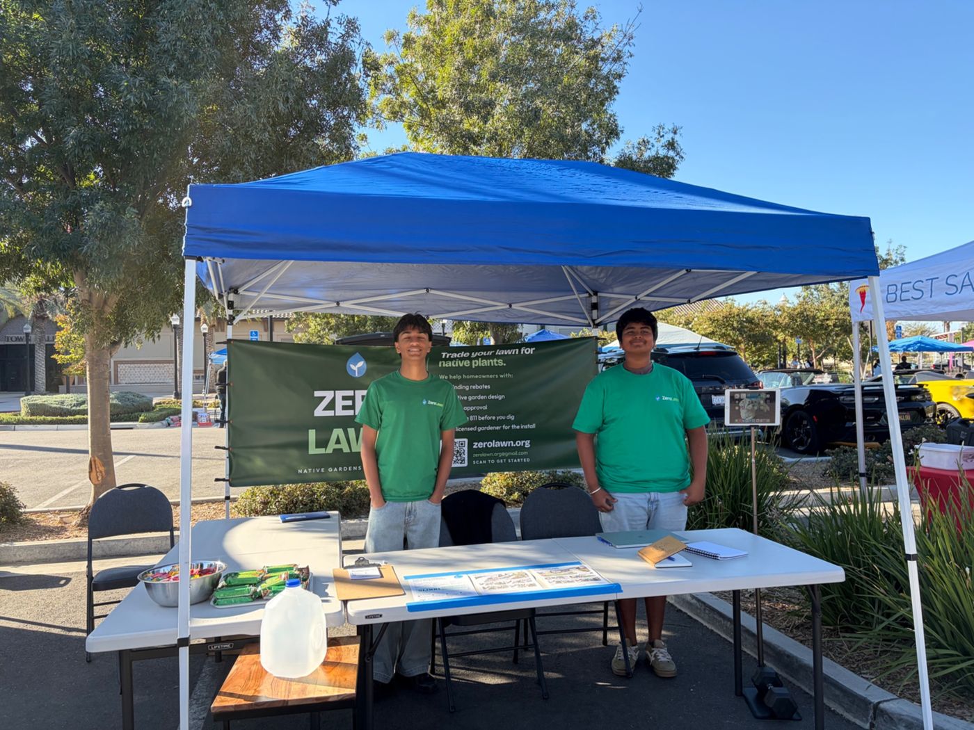 Two ZeroLawn students standing at their booth under a blue canopy at the farmers market